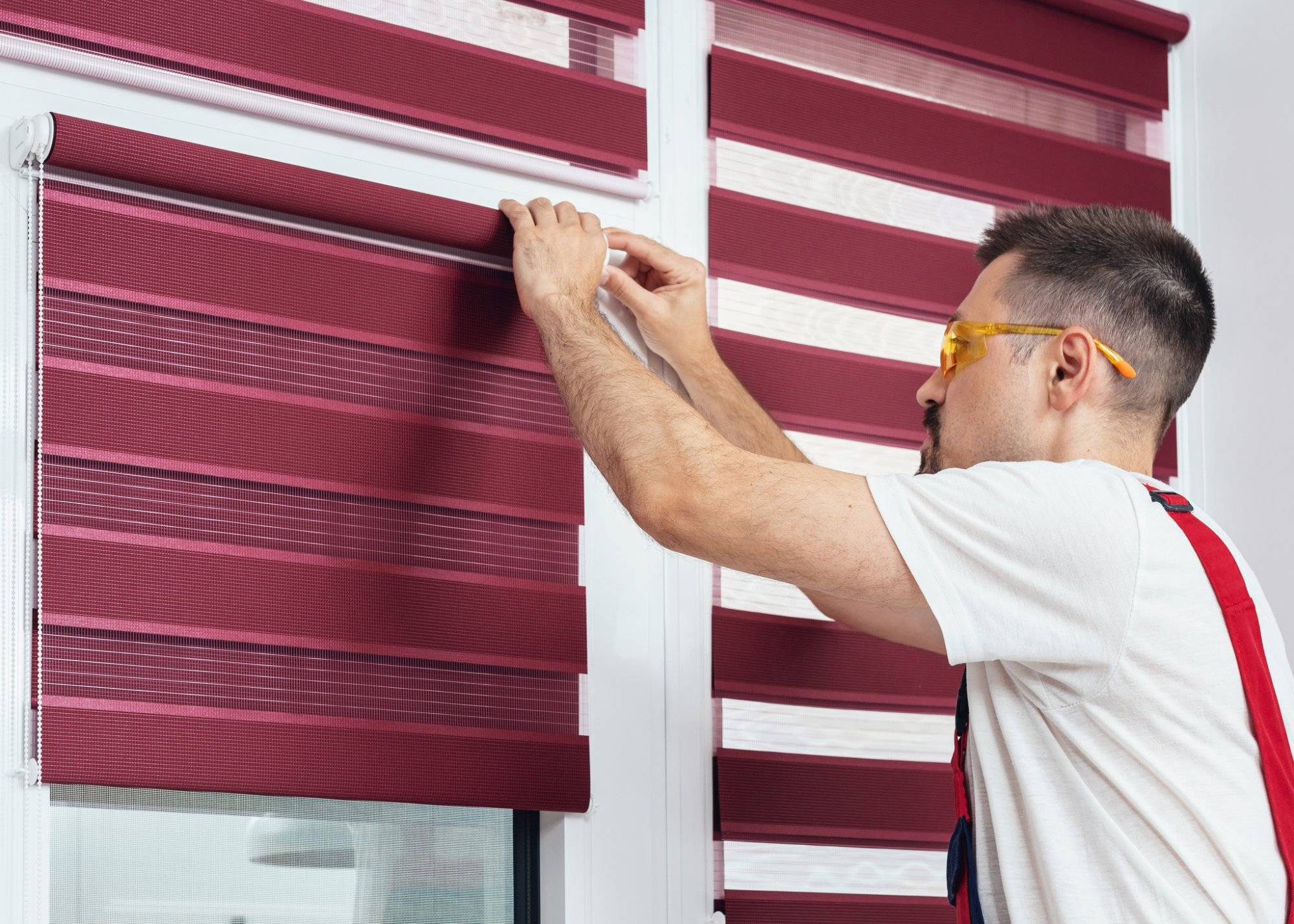 Young man in a uniform installing new window blinds Young man in a uniform installing new window blinds